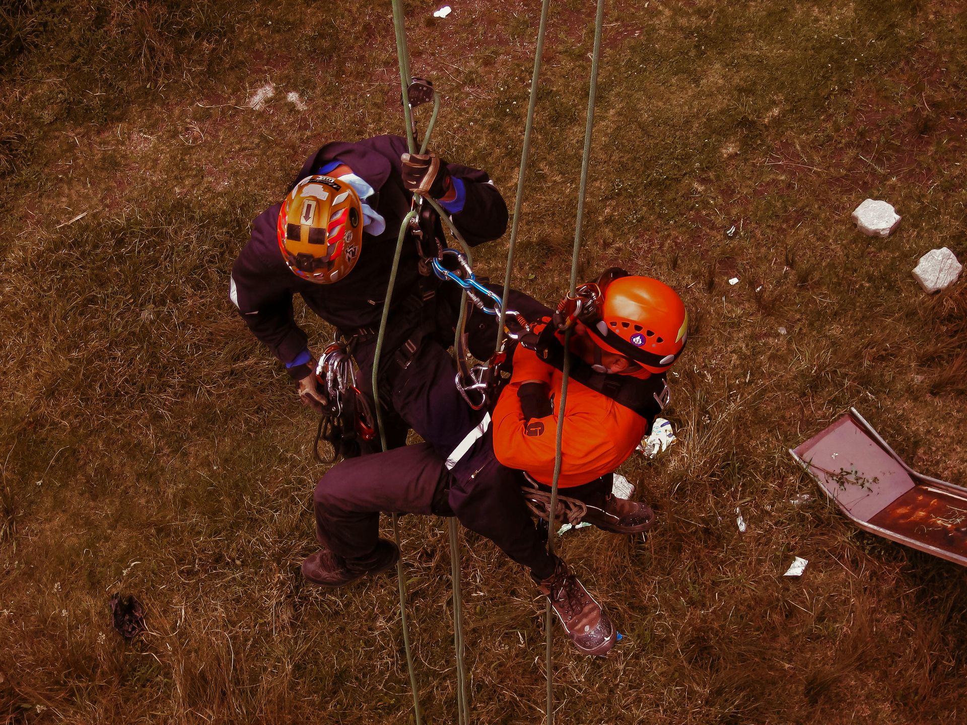 Two people in safety gear rappel down a grassy slope; one assists the other.