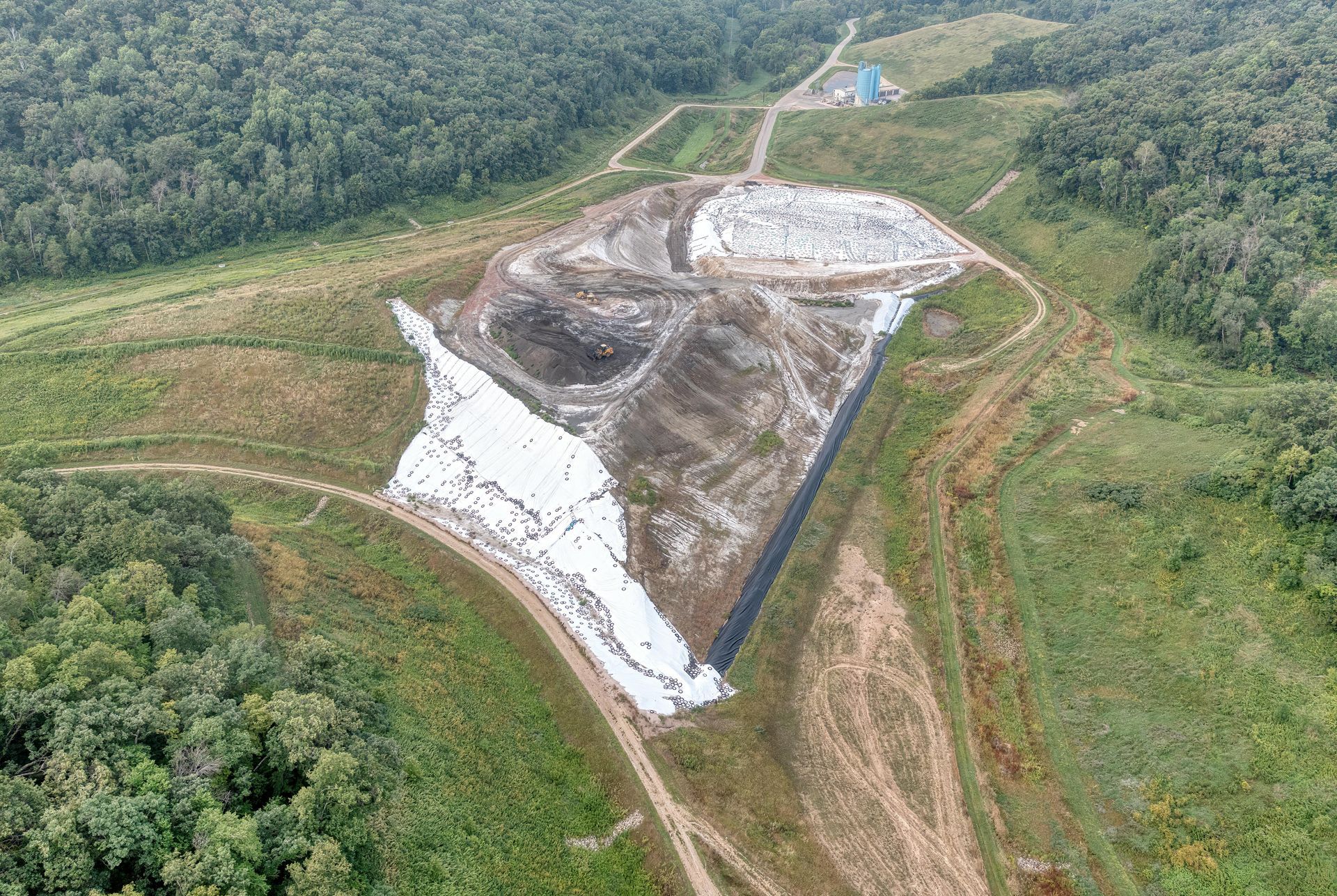 Aerial view of a landfill, partially covered in white sheeting, surrounded by cleared land and forest.