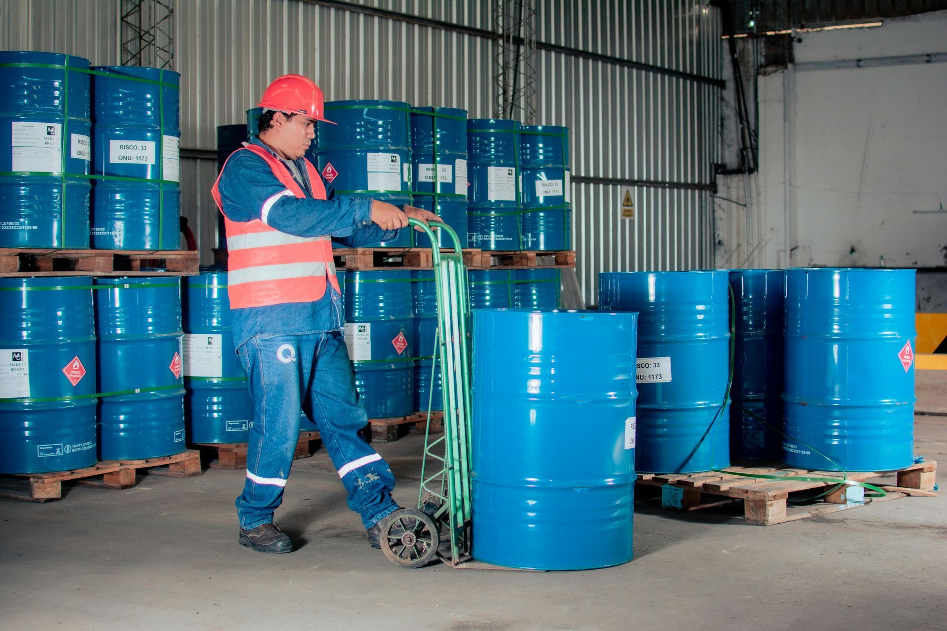 Warehouse worker using a hand truck to move a blue barrel. Pallets and other barrels in background.