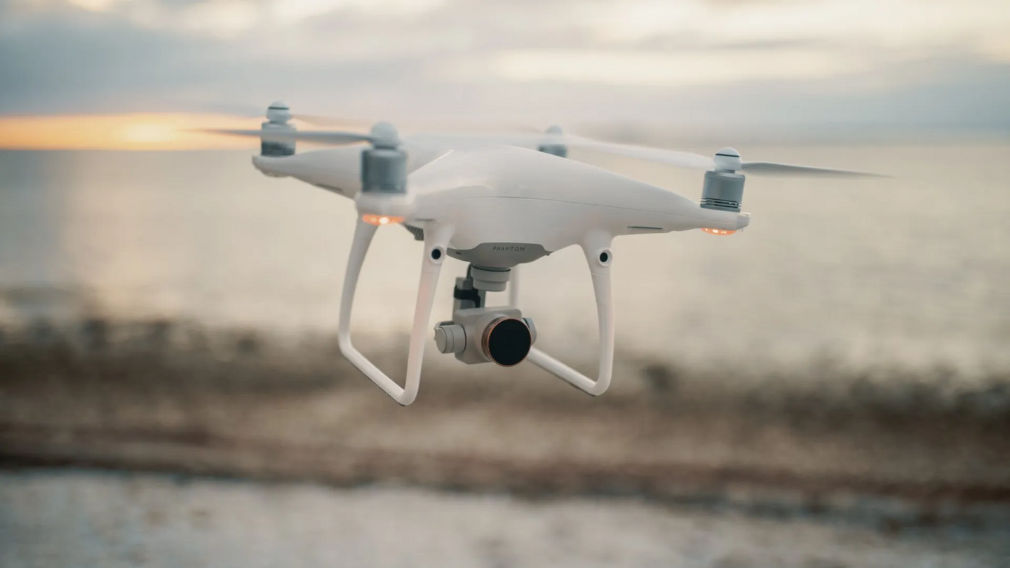 White drone hovering in air, camera angled down, blurred ocean background.