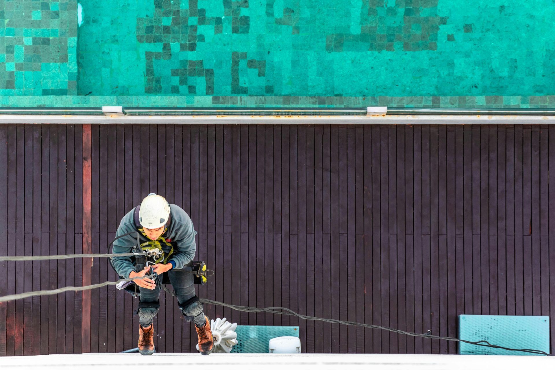 Person in rope access safety gear rappelling down a dark wooden wall; teal-green roof above.