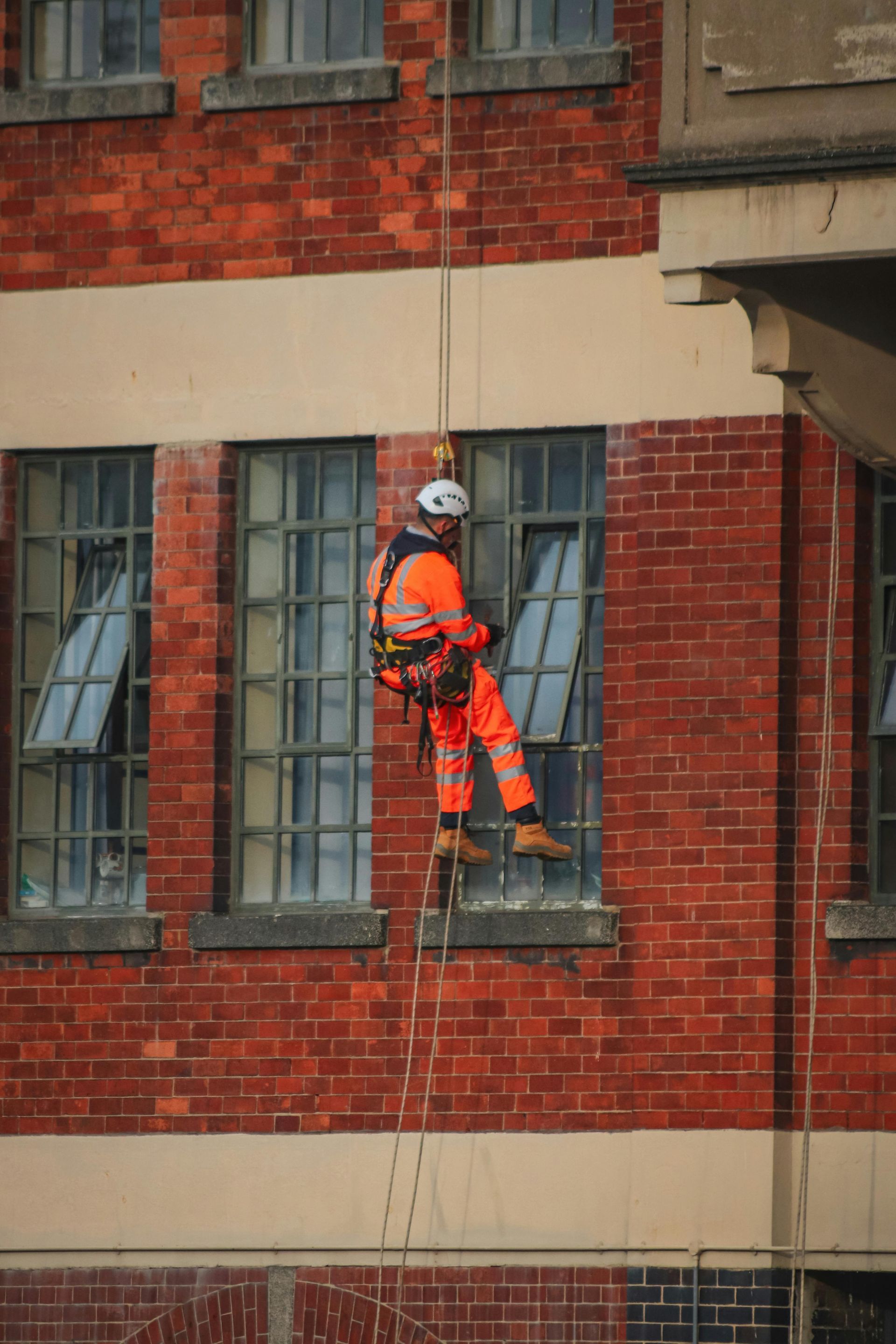 Rope access level 3 worker in orange suit, descending a red brick building, working on a window.