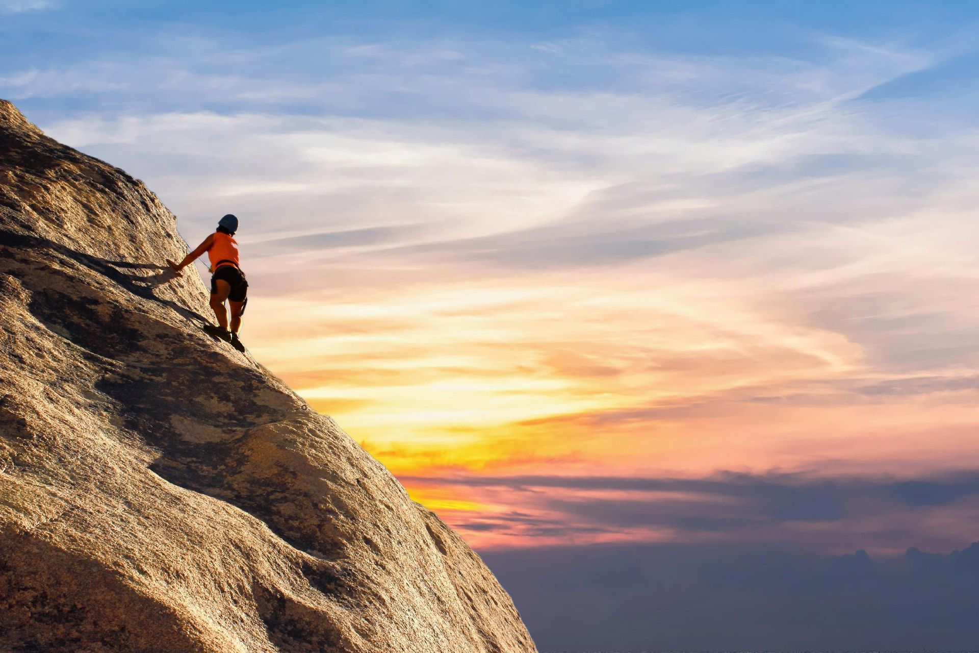 Rock climber ascends a steep rock face at sunset, silhouetted against colorful sky.