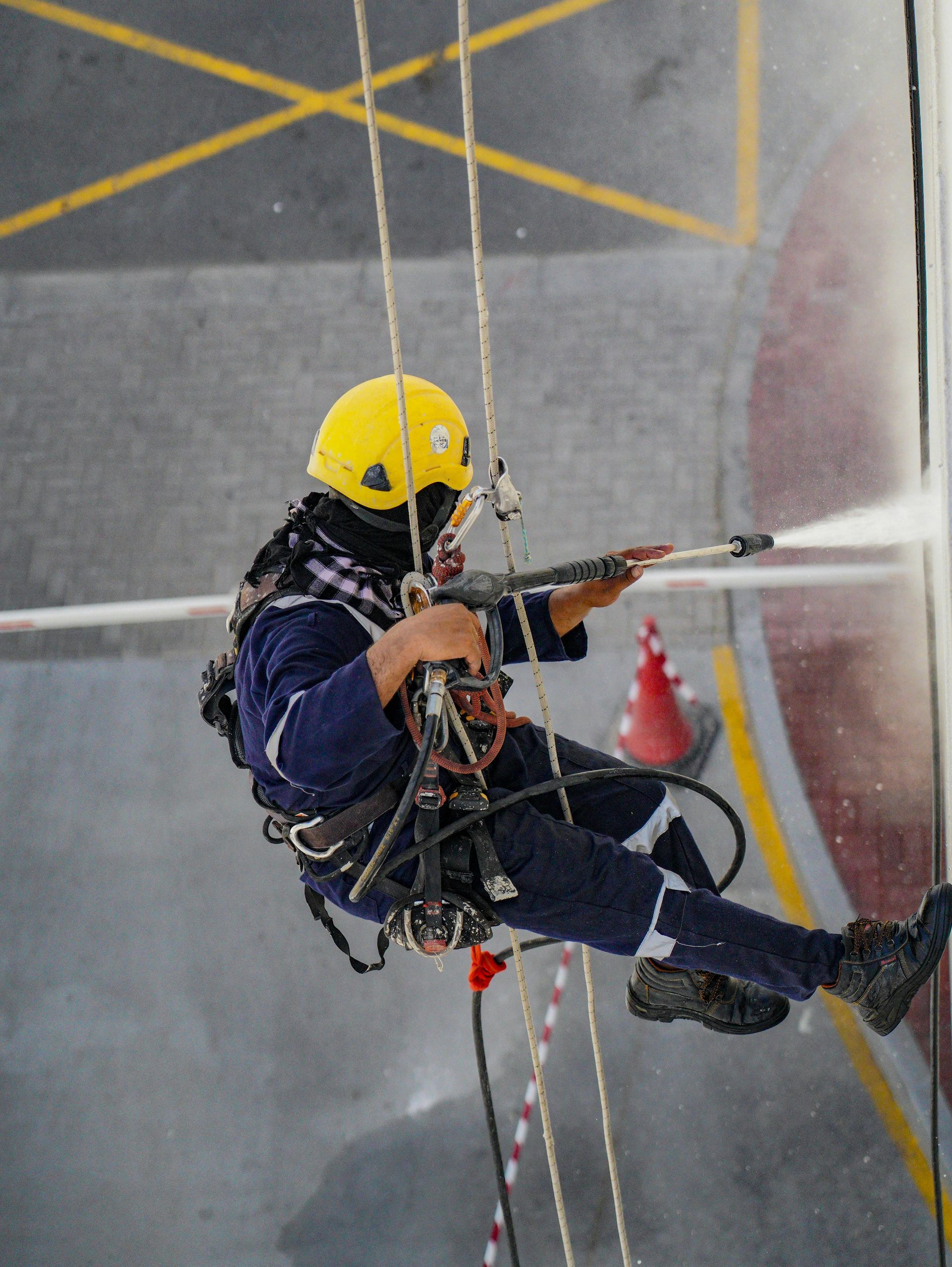 A man in a yellow helmet and harness is jet washing from ropes on a building