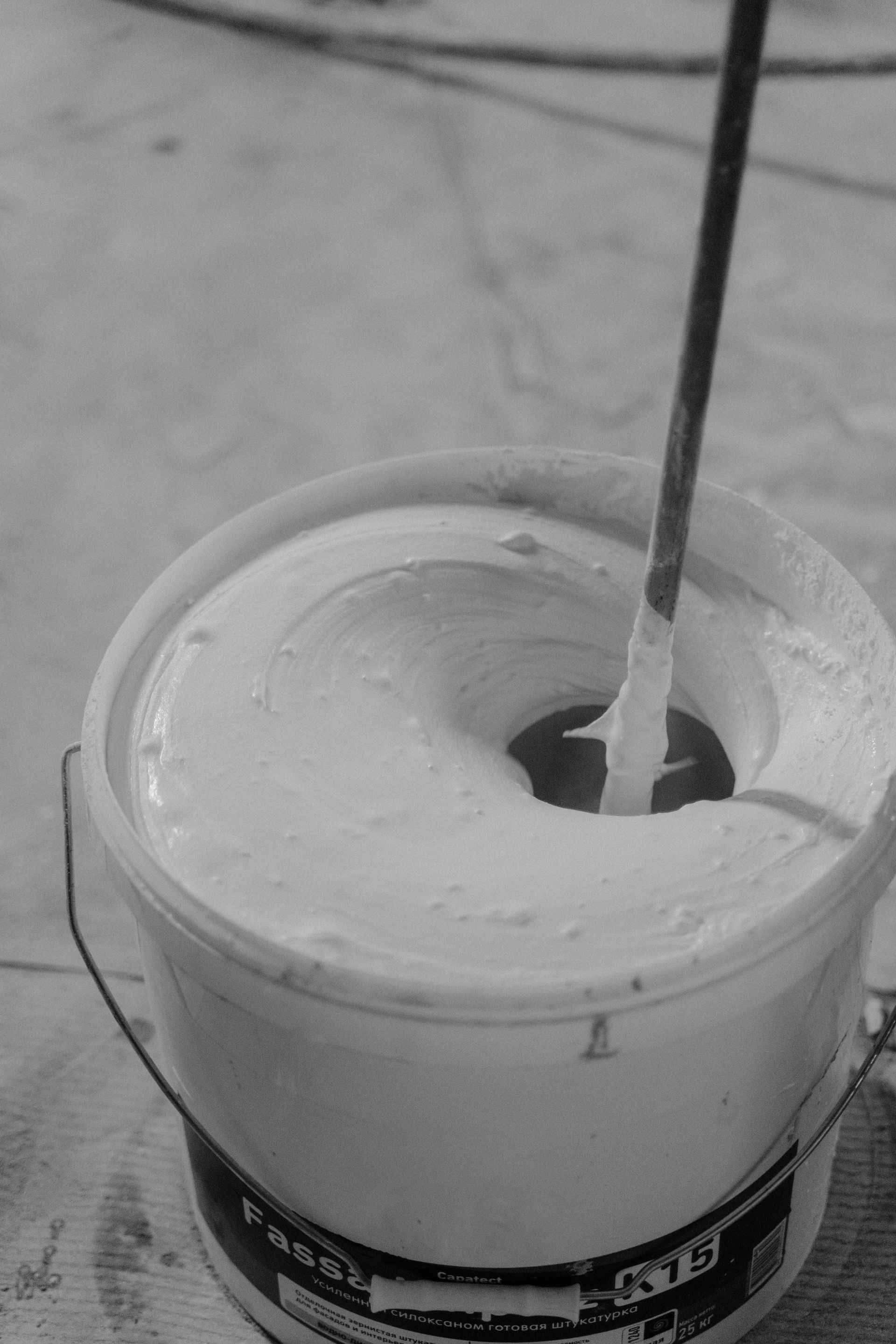 A black and white photo of a bucket of paint with a industrial paint paddle mixing in it.