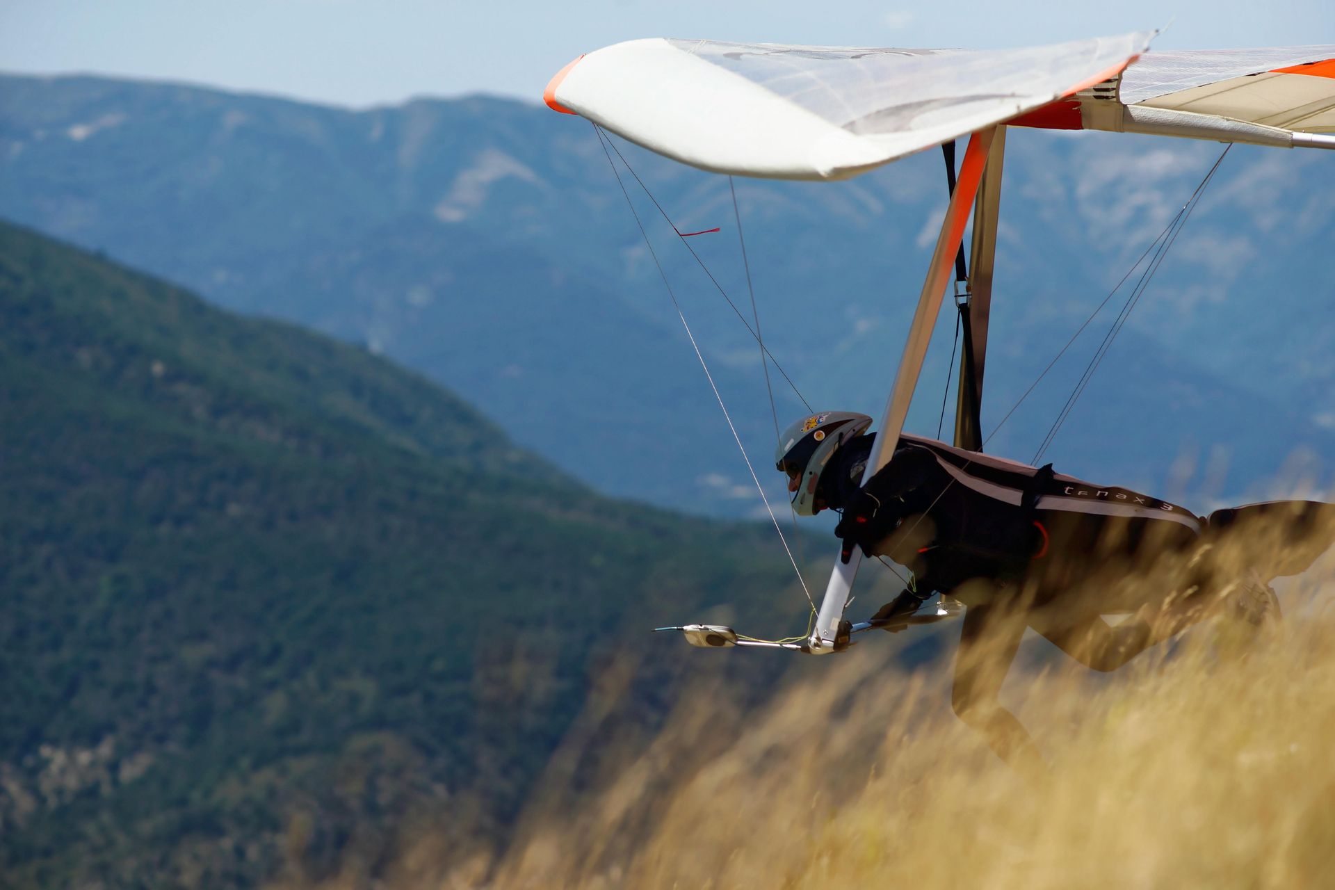 Hang glider pilot taking off from a grassy hilltop against a backdrop of mountains. The glider is white and orange.