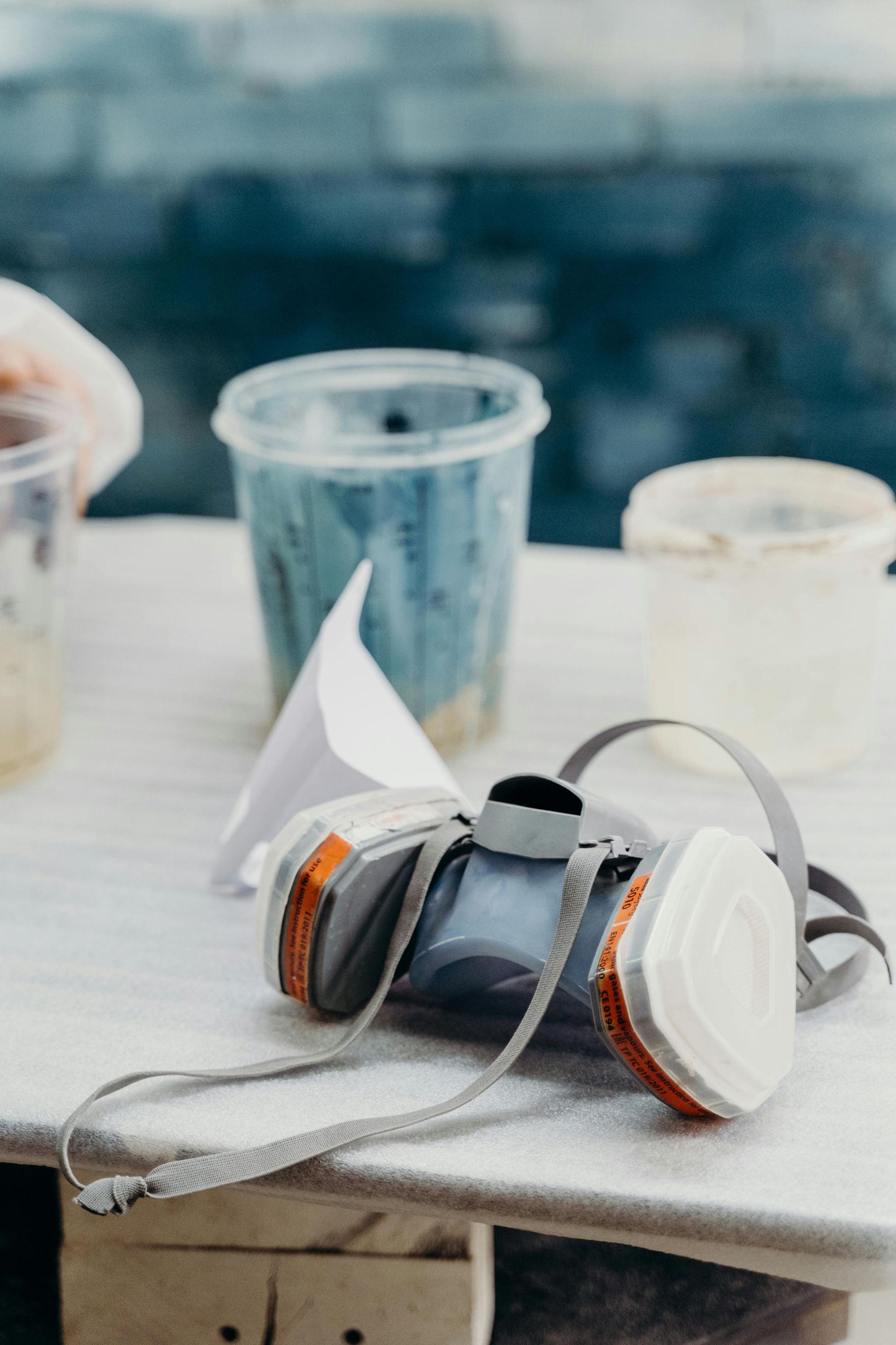 Respirator mask with filters, white plastic paint containers, and funnel on a work surface.