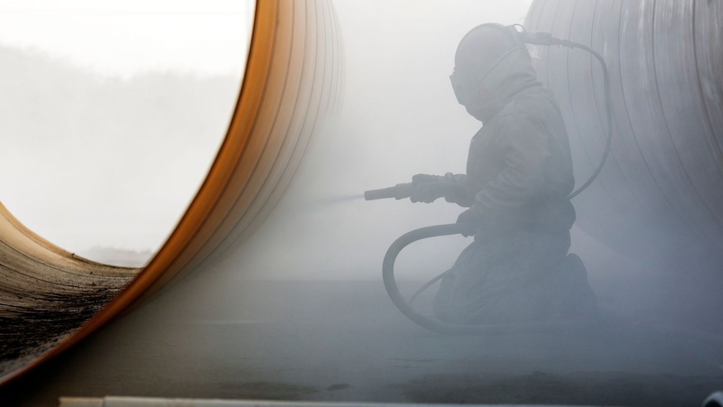 Worker in protective suit sandblasting large metal pipe, creating a cloud of dust.
