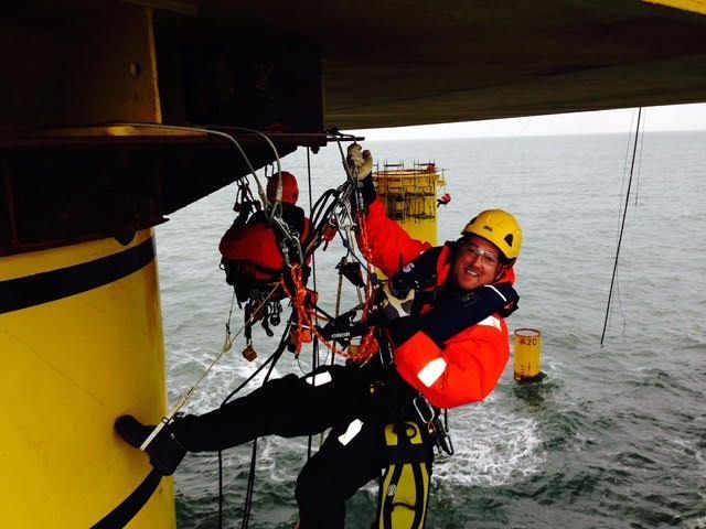 David in safety gear rappelling down a yellow offshore oil rig leg over the ocean. Smiling, orange jacket, yellow helmet.