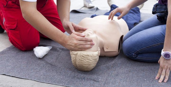 Two people practice CPR on a mannequin lying on a mat outdoors.