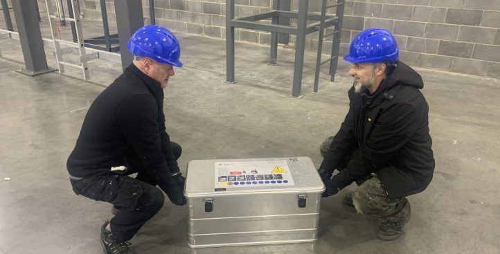Two men in blue hard hats lift a metal box in a concrete industrial space.