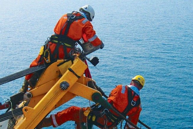 Workers in orange safety gear on a yellow offshore structure over the sea doing NDT inspections