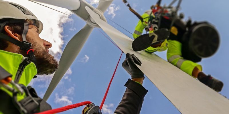 Rope access workers in safety gear on a wind turbine blade, one with a beard, another handling a rope.