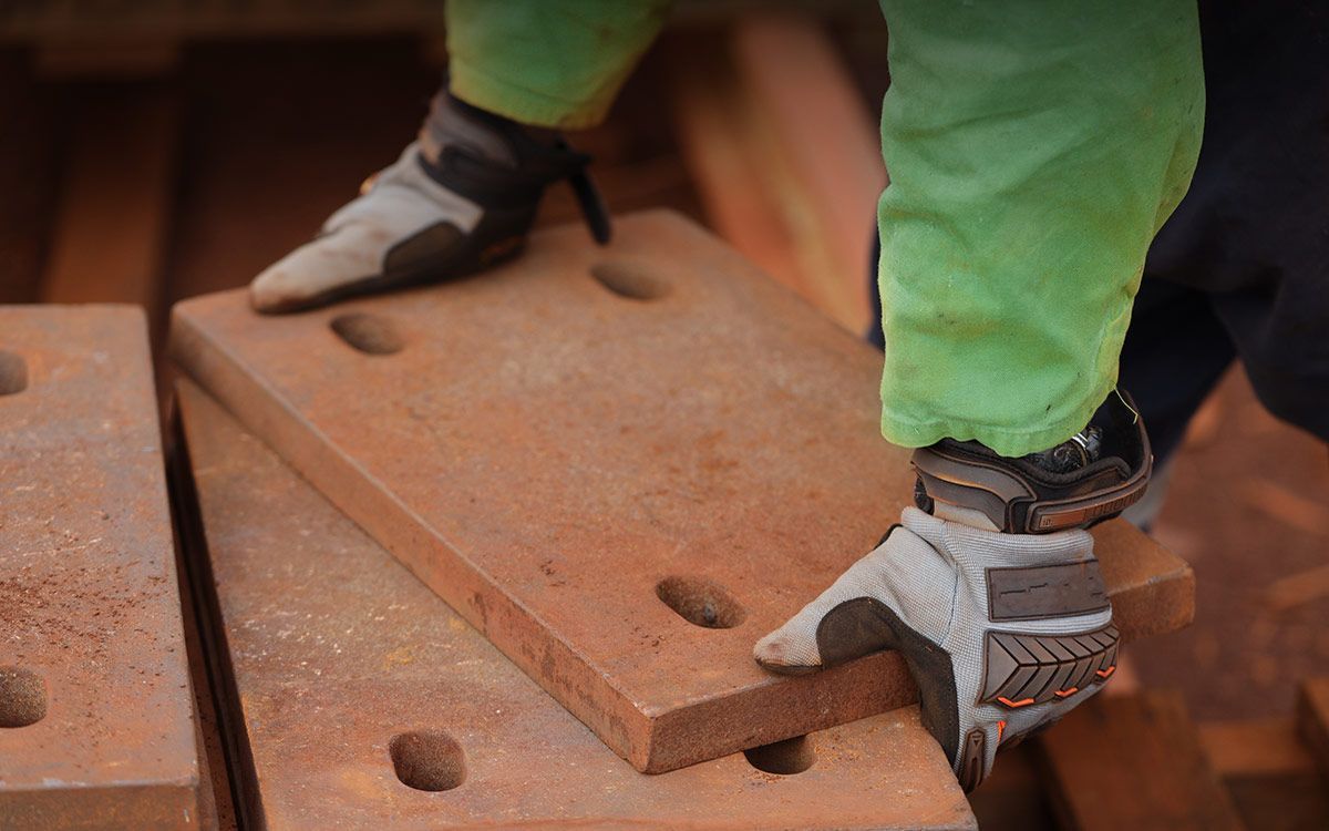 Worker placing a large, rusty metal plate. They wear gloves and green pants. Indoors, likely industrial setting.