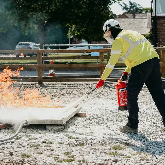 Person in safety gear extinguishing a fire with a fire extinguisher outdoors.