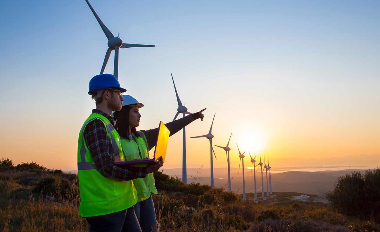 Two engineers at wind farm at sunset; one points, one holds laptop.
