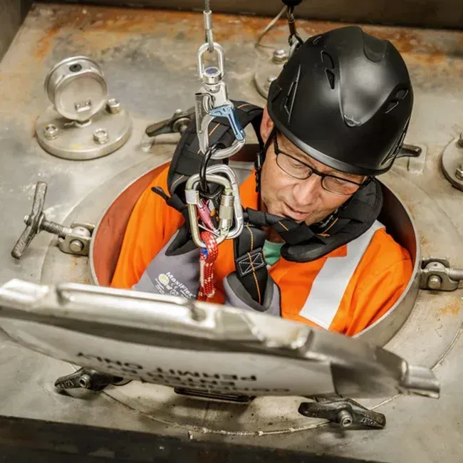 Worker in orange vest and helmet entering tank, secured by harness.