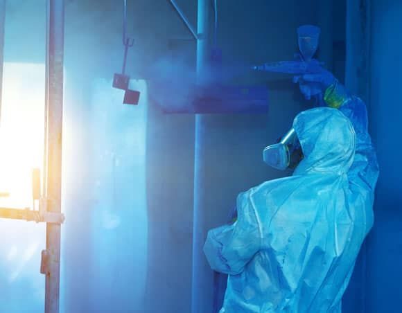 Person in protective suit spray painting objects in a blue booth.