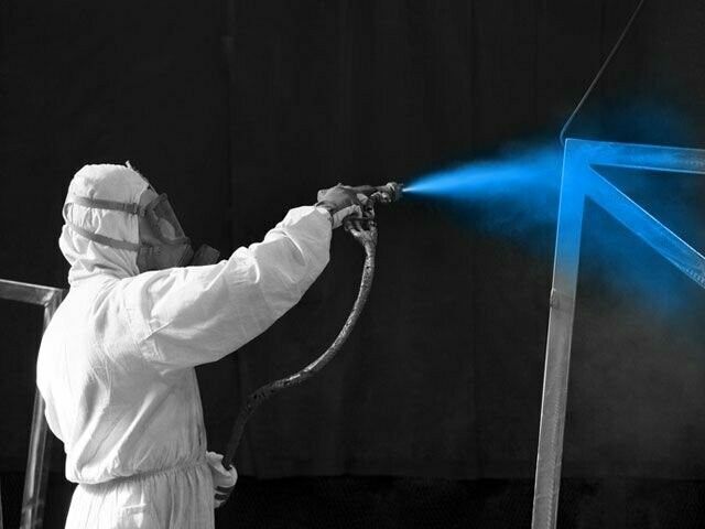 Person in protective suit spray-painting a metal frame with blue paint.