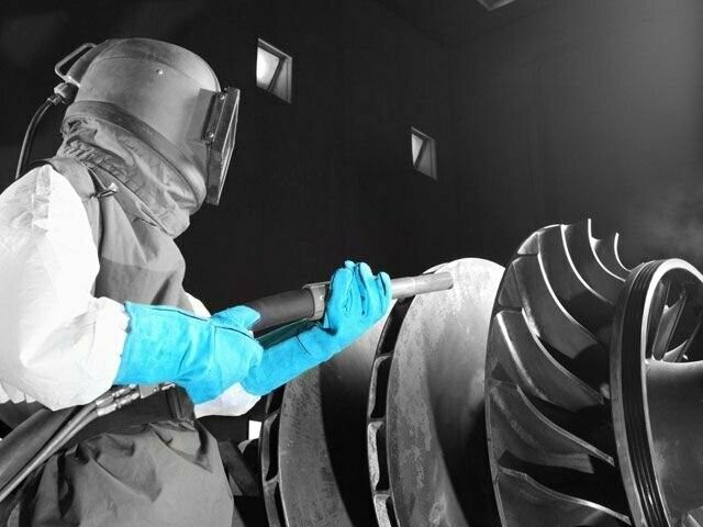 Person in protective gear sandblasting a turbine wheel; monochrome setting with blue gloves.
