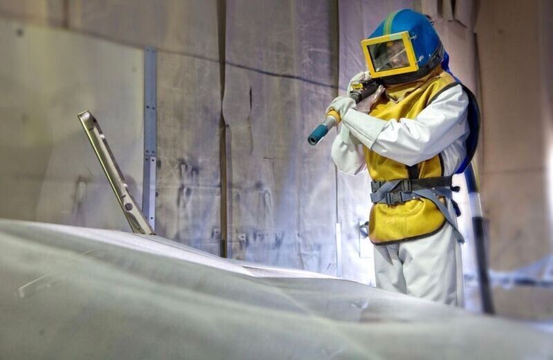 Person in protective gear sandblasting a surface in an industrial setting.