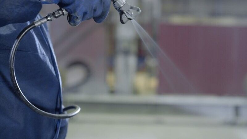 Person in blue coveralls spraying paint with a paint sprayer.