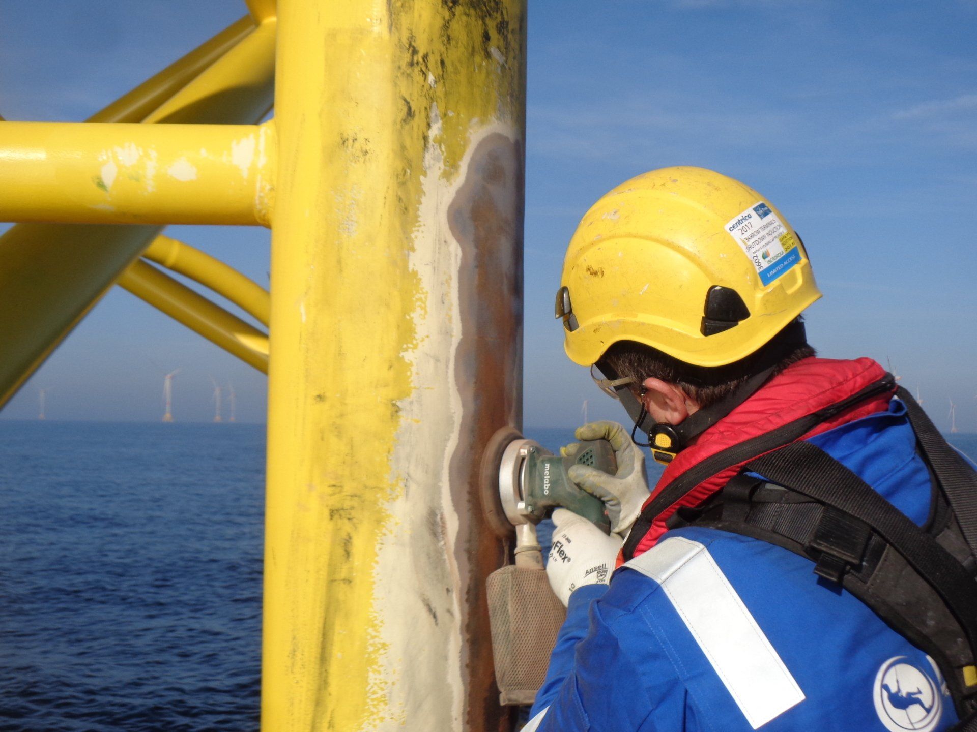 Worker in blue Dangle coveralls and a yellow helmet sanding a yellow offshore structure, with the sea and wind turbines
