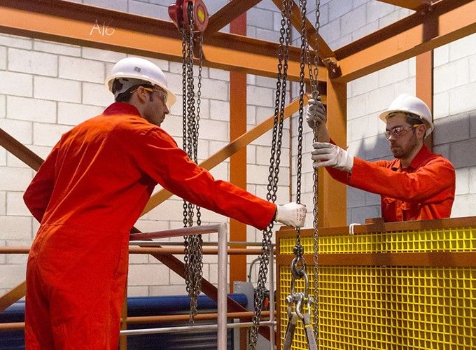 Two workers in orange coveralls and white hard hats use chains to lift something in an industrial setting.