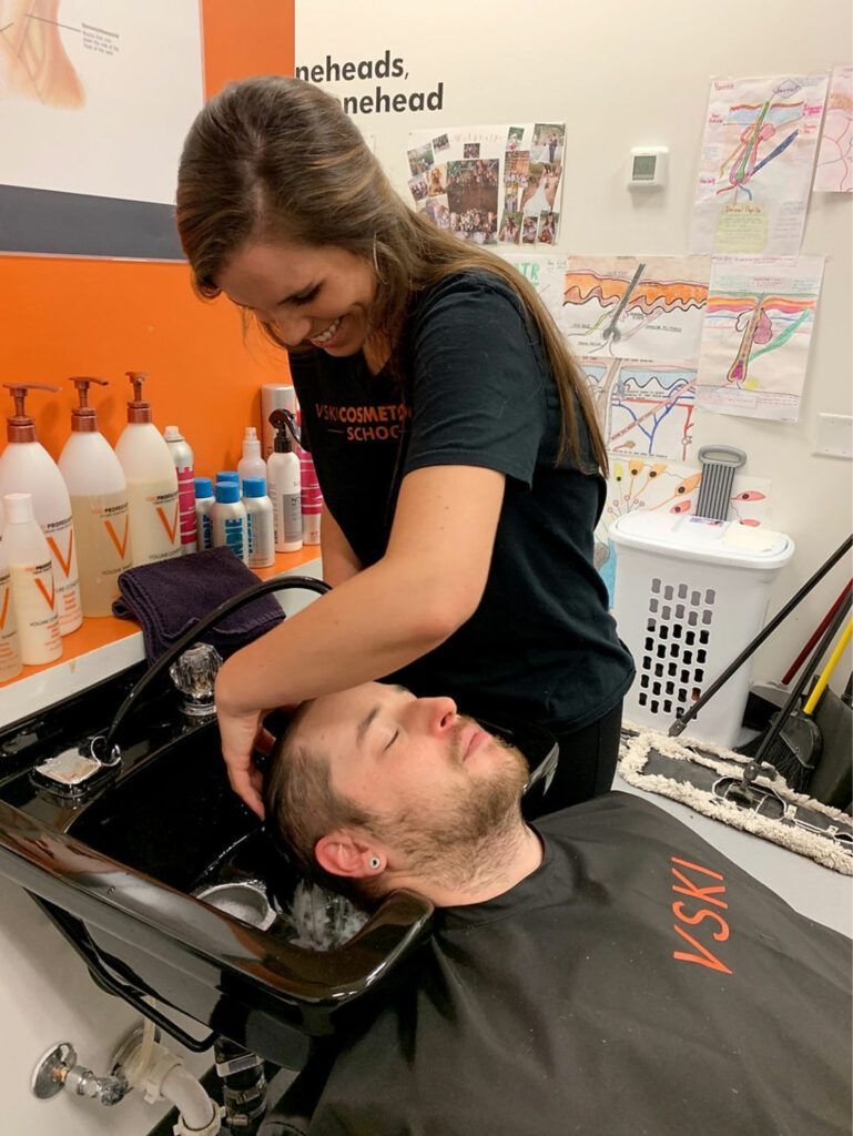 Hairdresser washing a man's hair in a salon sink, with product bottles and posters in the background.