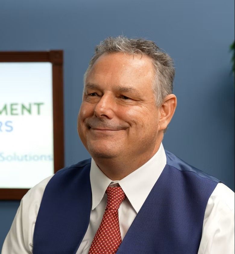 Man in white shirt, blue vest, red tie, smiling. Office setting with blue wall.