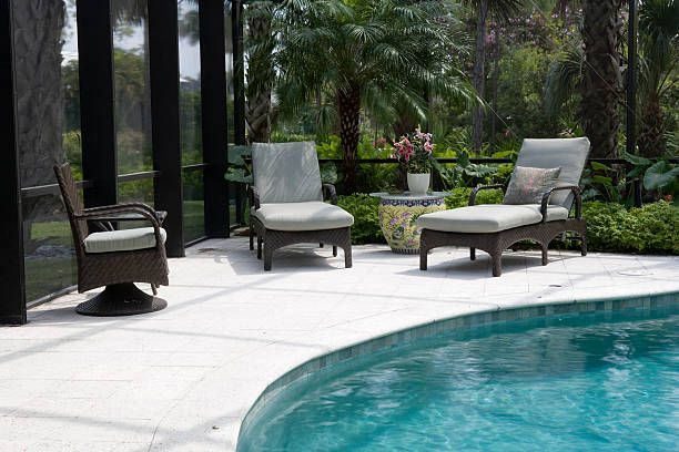 Outdoor seating area with pool. Three wicker chairs with cushions on a white patio. Green plants and a blue pool.