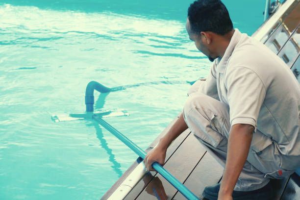 Man cleaning a swimming pool with a long-handled vacuum, blue water and a wooden deck.