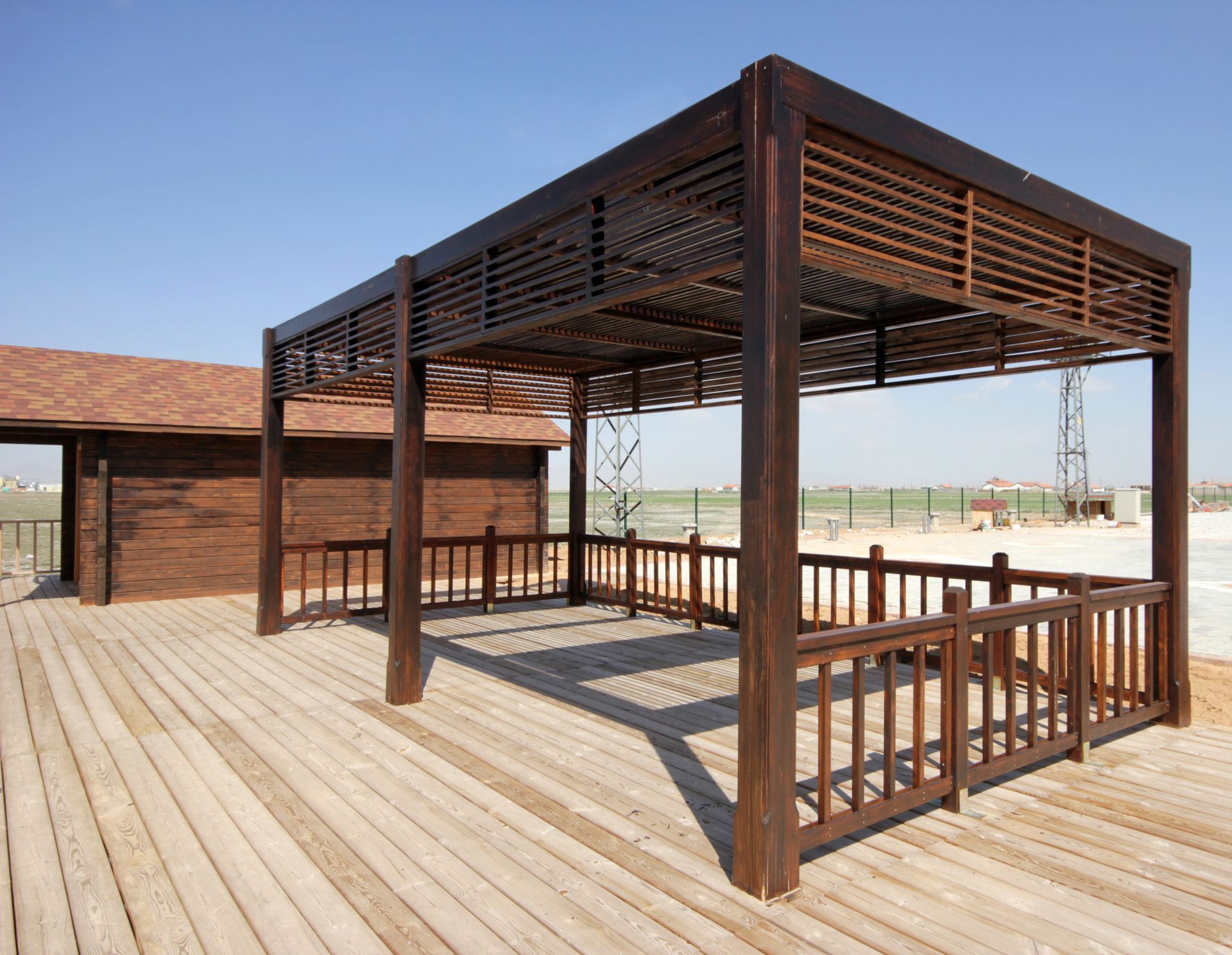 Wooden gazebo on a wooden deck with railing. Brown building in the background. Sunny day.
