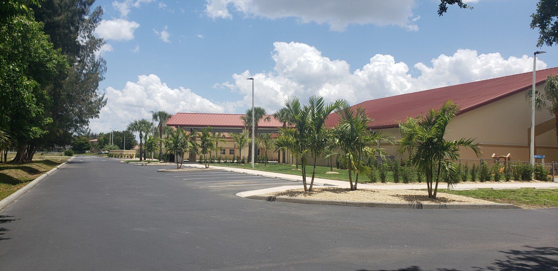 An asphalt road leads to a tan building with a red roof, palm trees, and a bright blue sky.