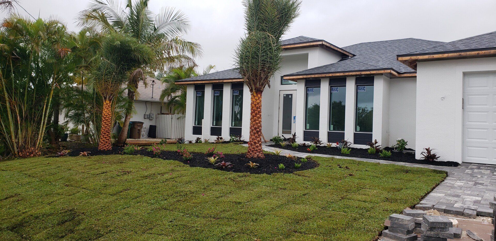 White house with large windows, palm trees, and fresh grass. Gray paved walkway. Overcast day.