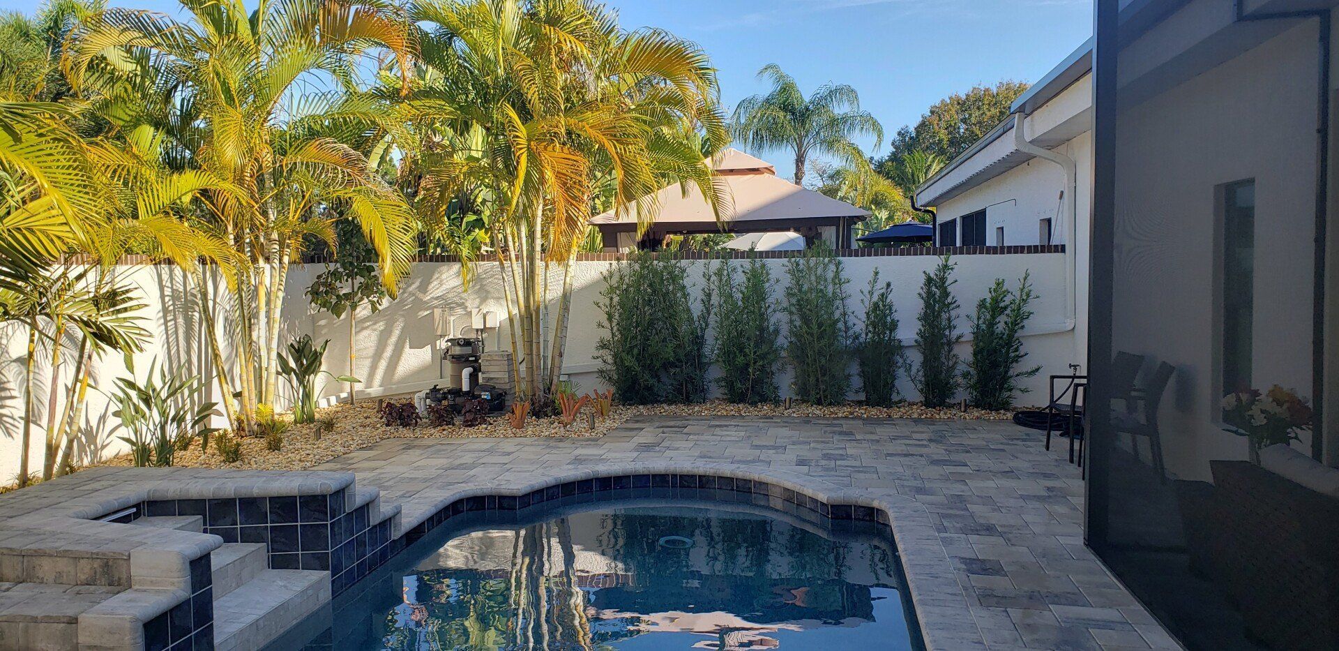 Backyard with pool, landscaping, white fence, and a gazebo in the distance under a blue sky.