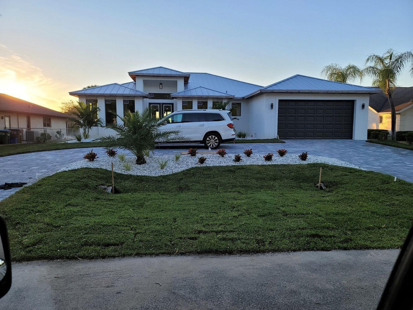 White house with a white SUV in the driveway, surrounded by a grassy lawn and landscaping, blue sky.