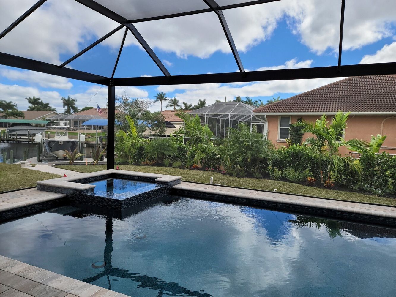 Swimming pool with adjacent hot tub under a screened enclosure; blue water reflects sky and clouds.