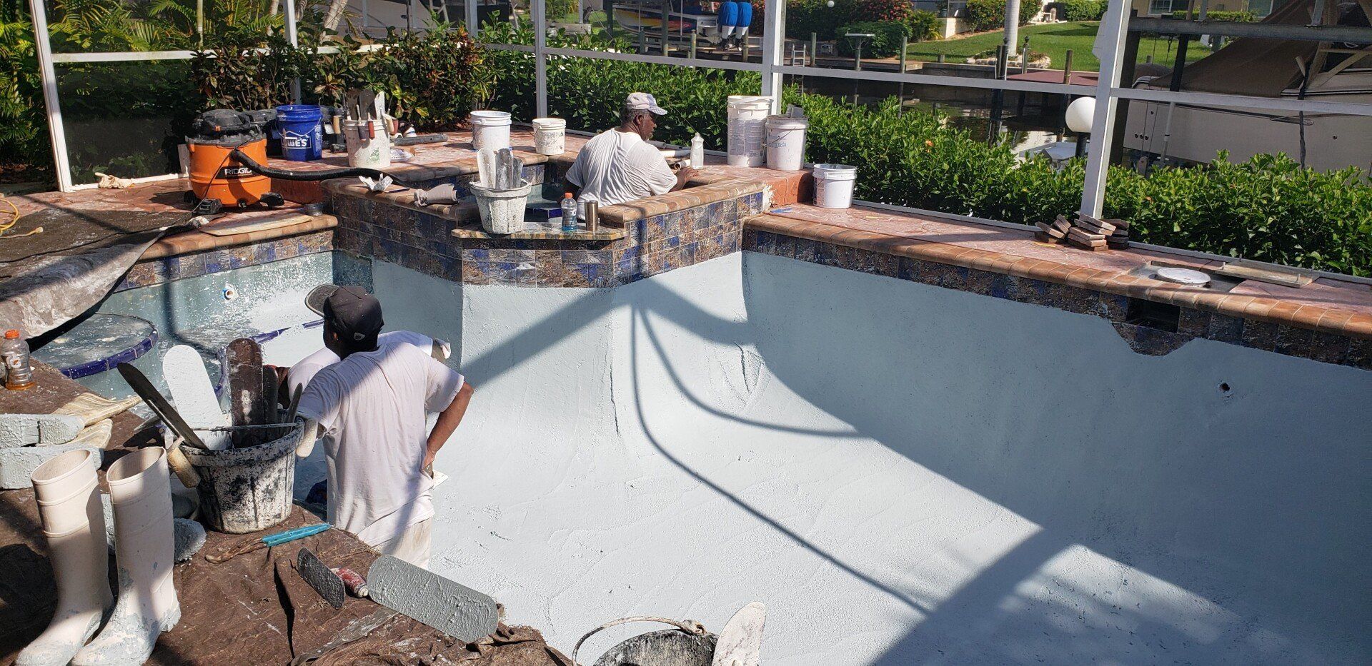 Men repainting a swimming pool with white paint, various tools and supplies visible.