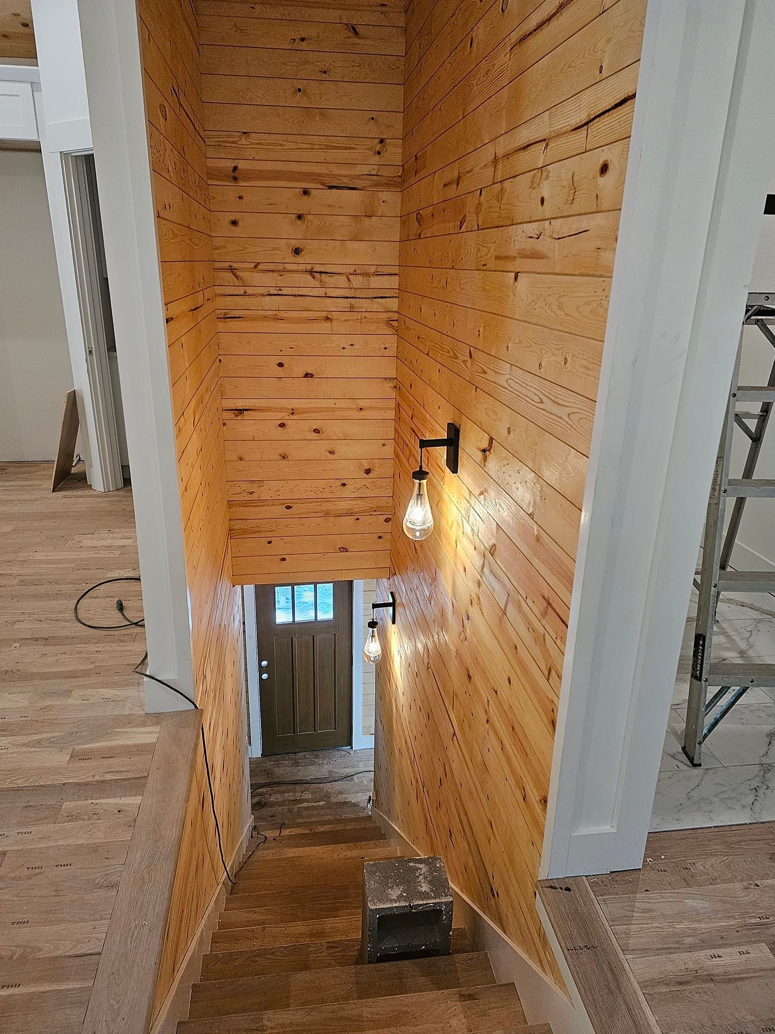 Staircase with wood-paneled walls and sconce lighting, leading down to a doorway. A ladder and unfinished flooring are visible.