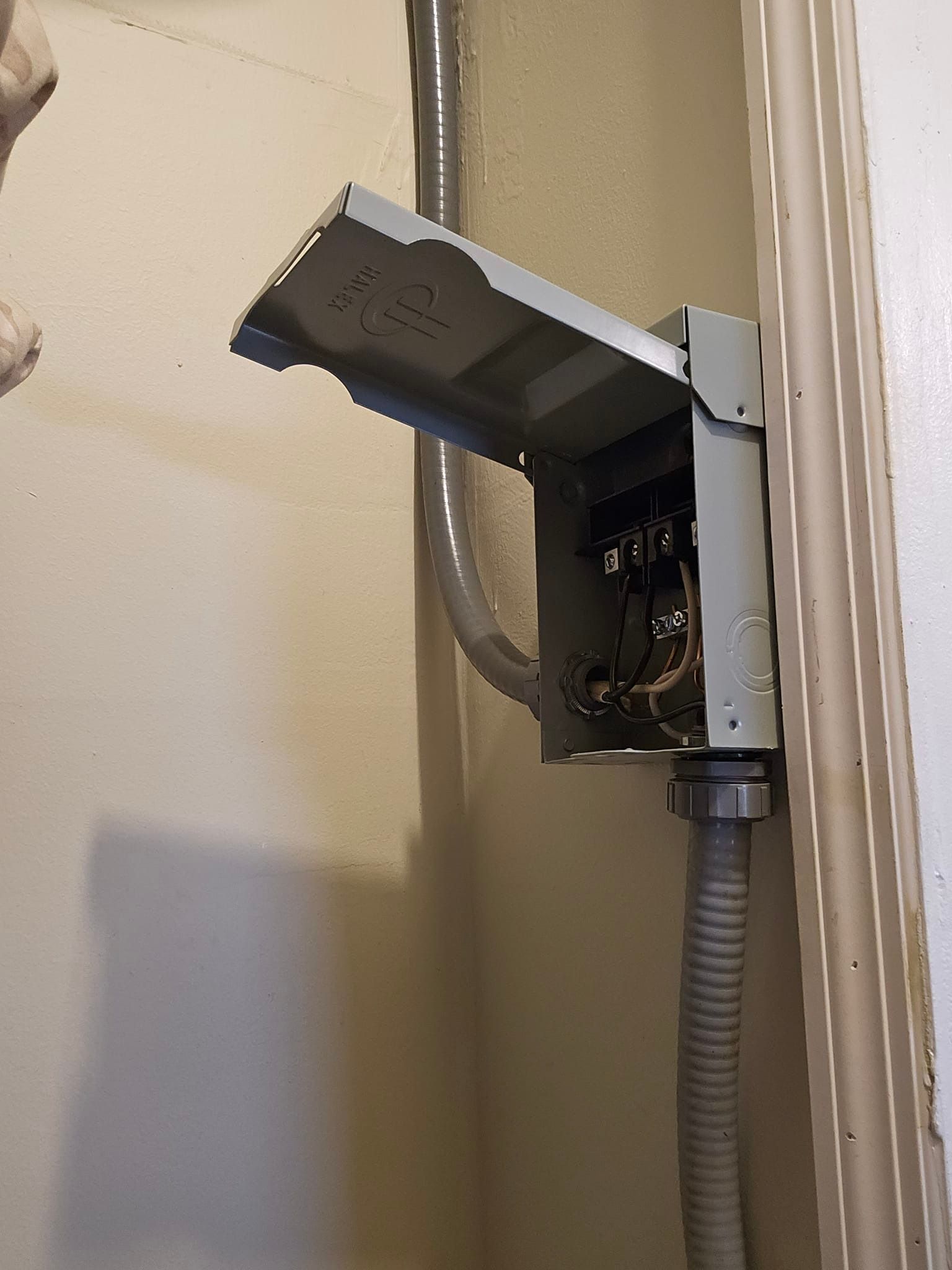 Open electrical box with wires, metal conduit, and gray flexible tubing on a beige wall.