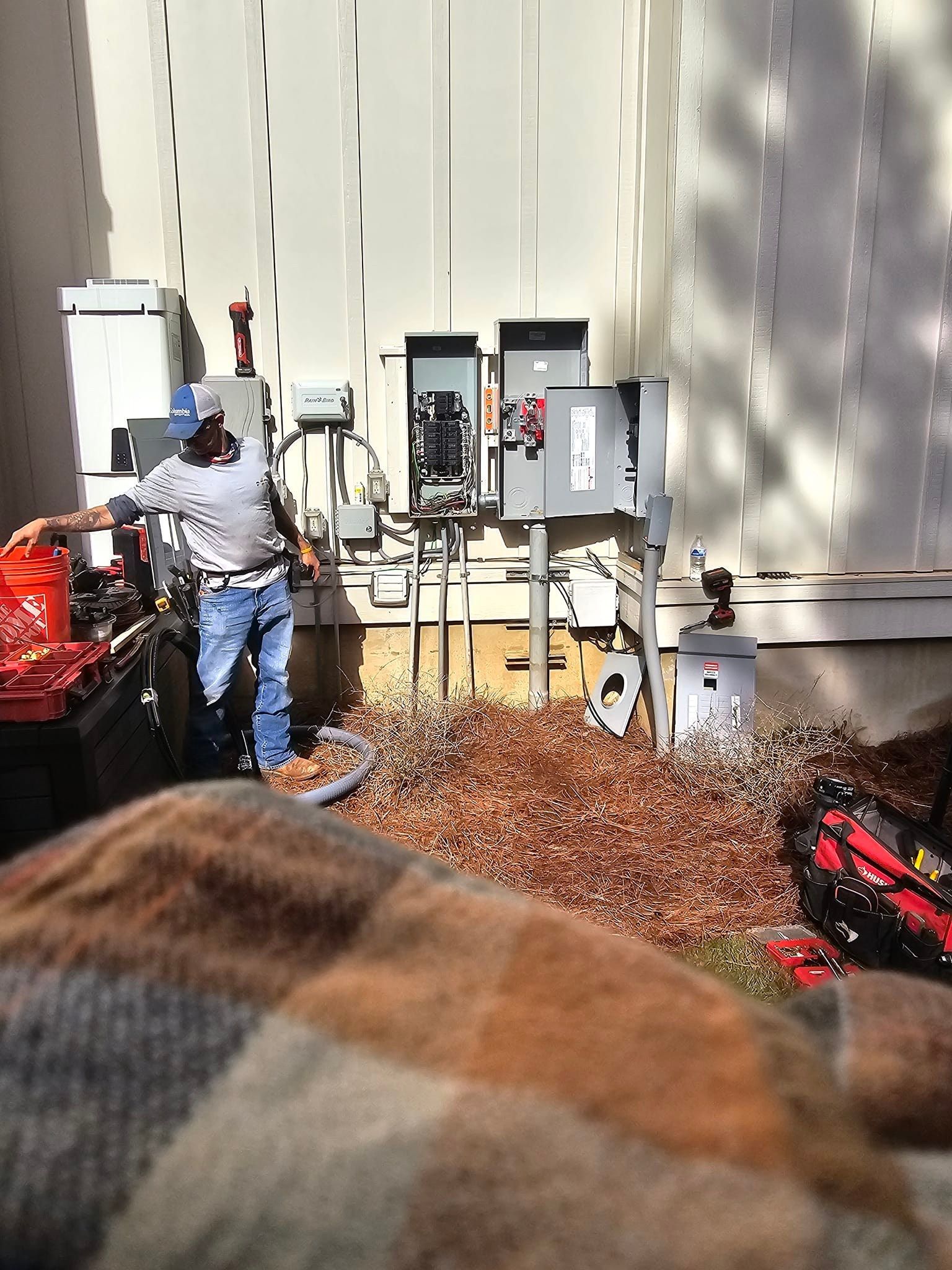 Man working on electrical boxes outside a building, surrounded by wood shavings.