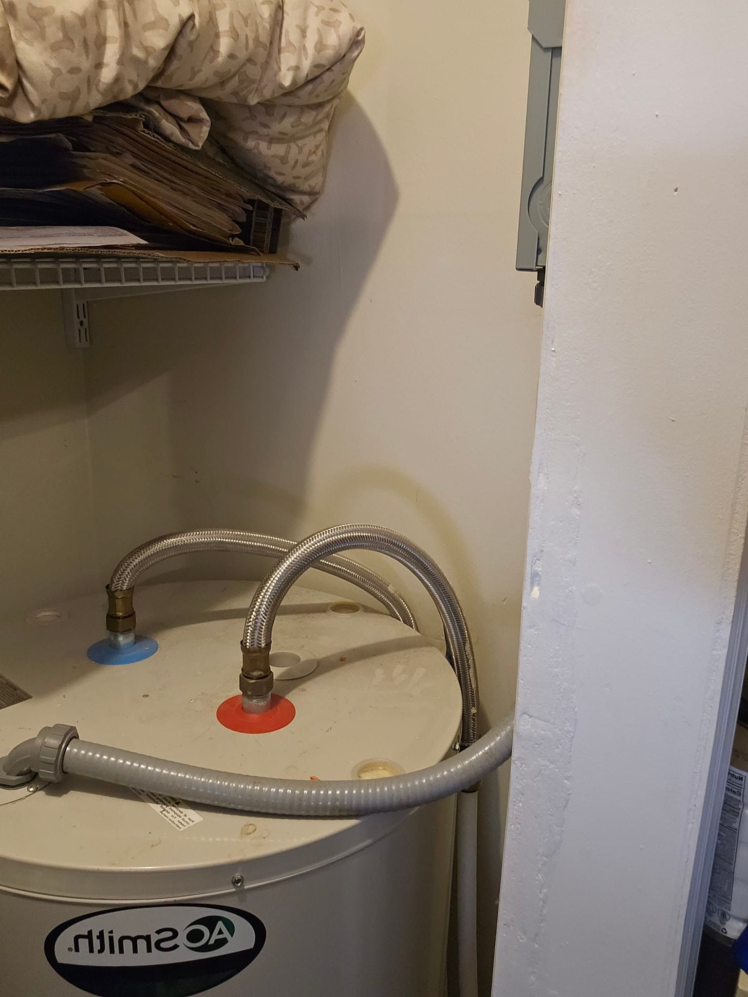 Water heater with flexible metal pipes connected, in a utility closet with a shelf. Red and blue valves visible.