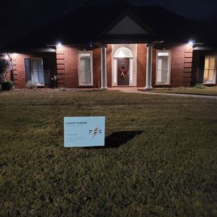 A sign on the lawn of a brick house at night. Lights illuminate the front door.