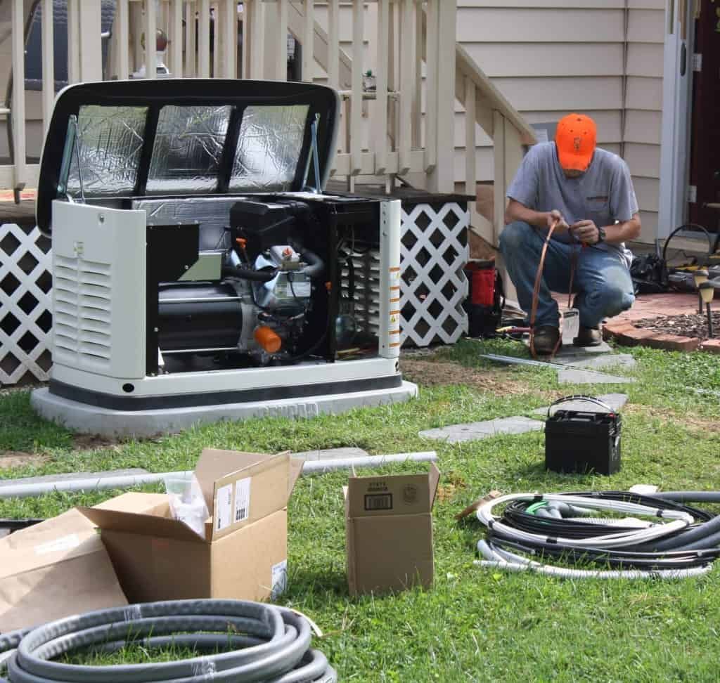Man installing generator on grass. Open generator, boxes, and wires nearby.