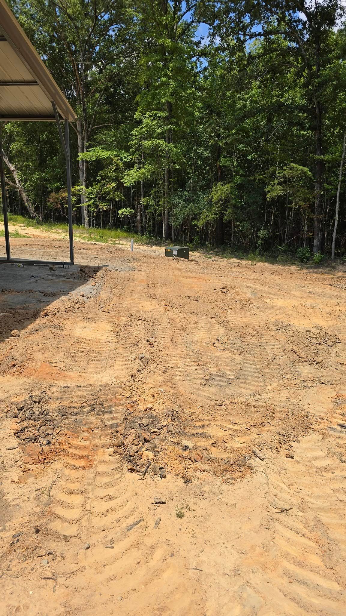 Dirt path in front of a metal-roofed structure, leading to a treeline. Sunny day.