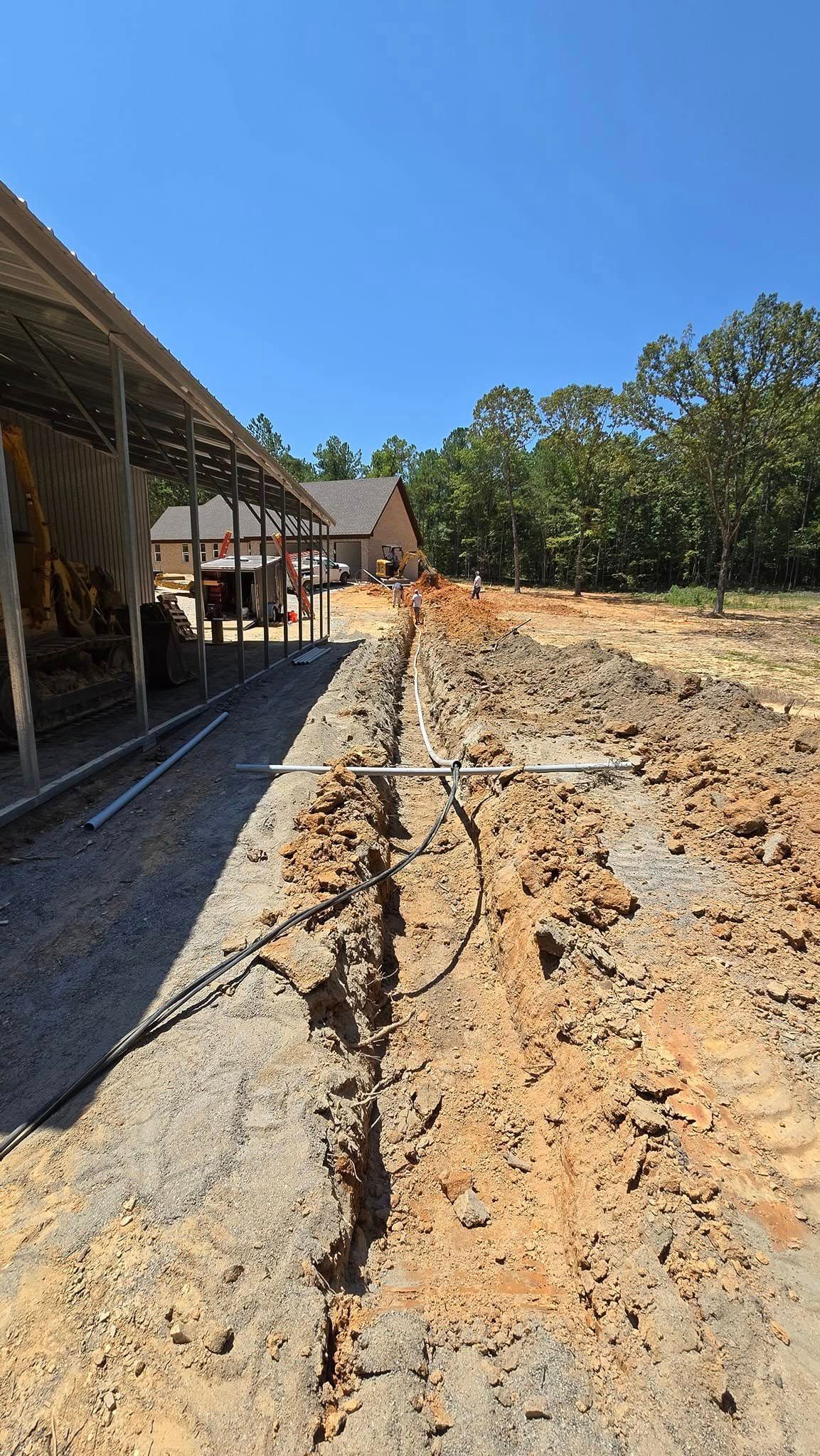 Construction site; trench dug in dirt, gravel path. Building with covered walkway on the left, trees and sky in the background.