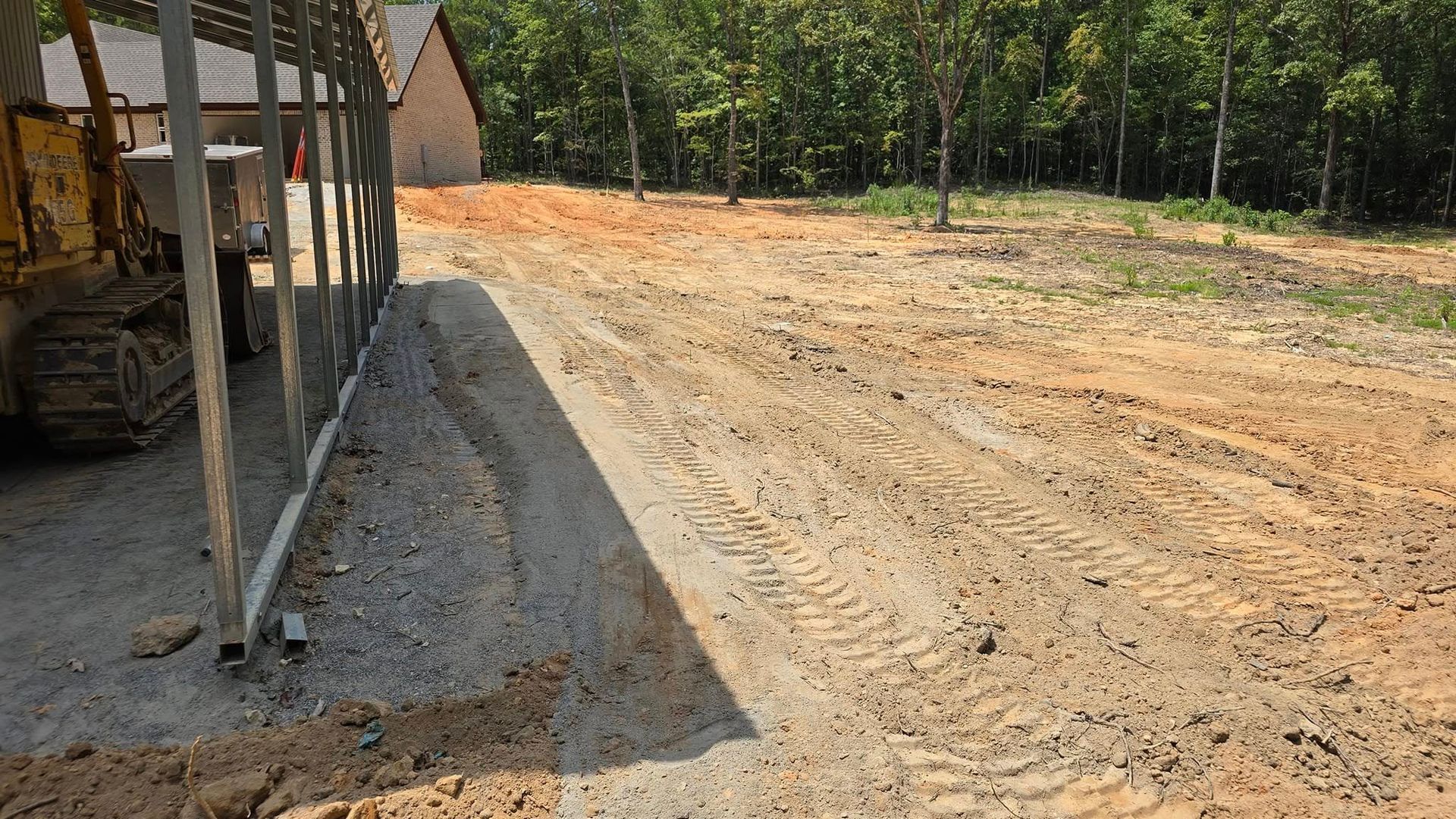 Dirt road with tire tracks, construction equipment, and a metal fence. Trees and a building in the background.