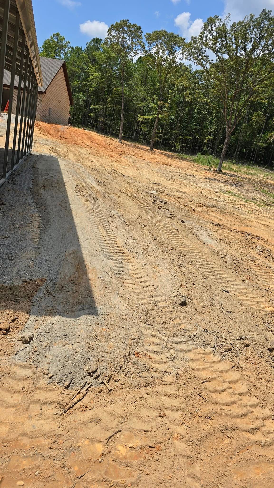 Dirt driveway with construction equipment tracks next to a building under construction, trees in background.