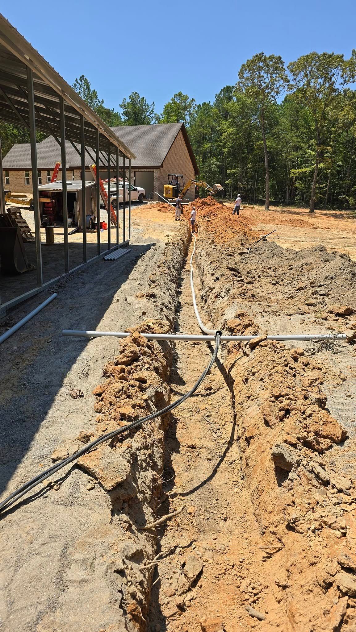 Trench dug in dirt, with conduit and wires visible. Buildings and trees in background under a blue sky.